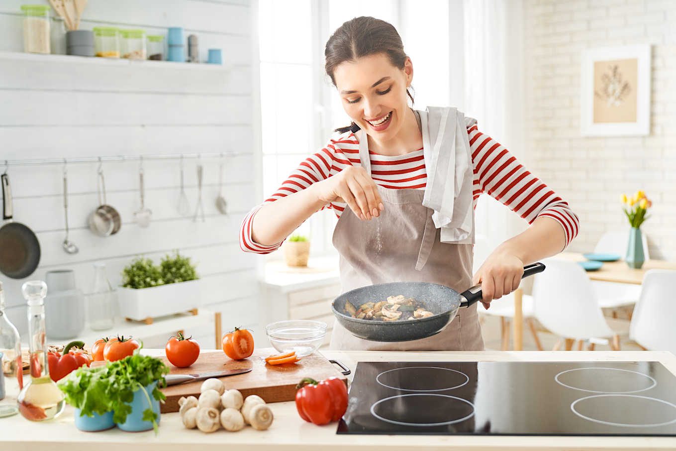 Woman cooking with a frying pan in a bright kitchen, surrounded by fresh vegetables.