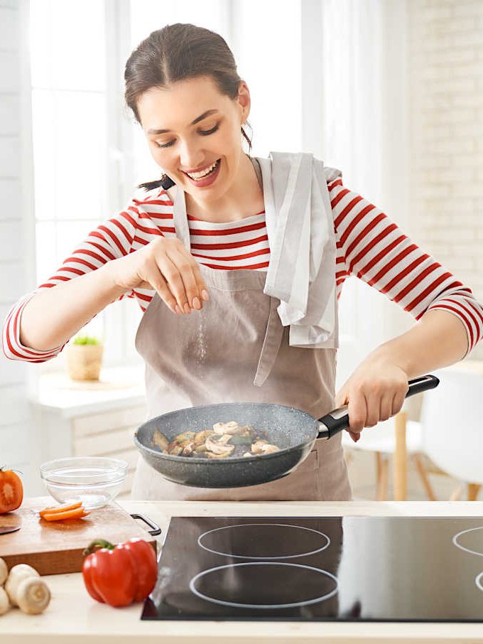 A woman in a striped shirt and apron sprinkles seasoning while cooking in a frying pan.