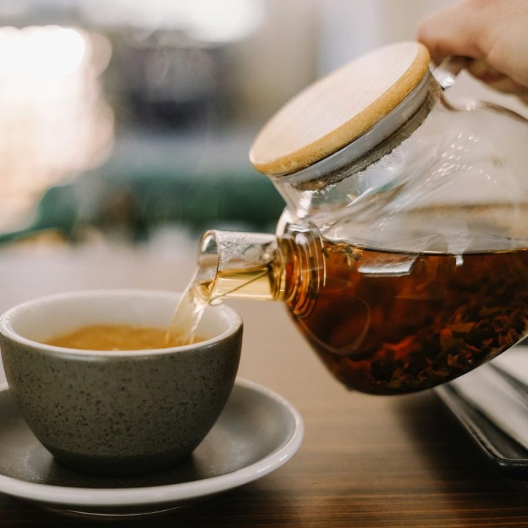 A person pours tea from a glass teapot into a gray cup on a wooden table.