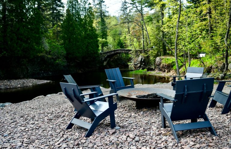 Chairs are arranged around a fire pit by a serene riverbank in a forested area.