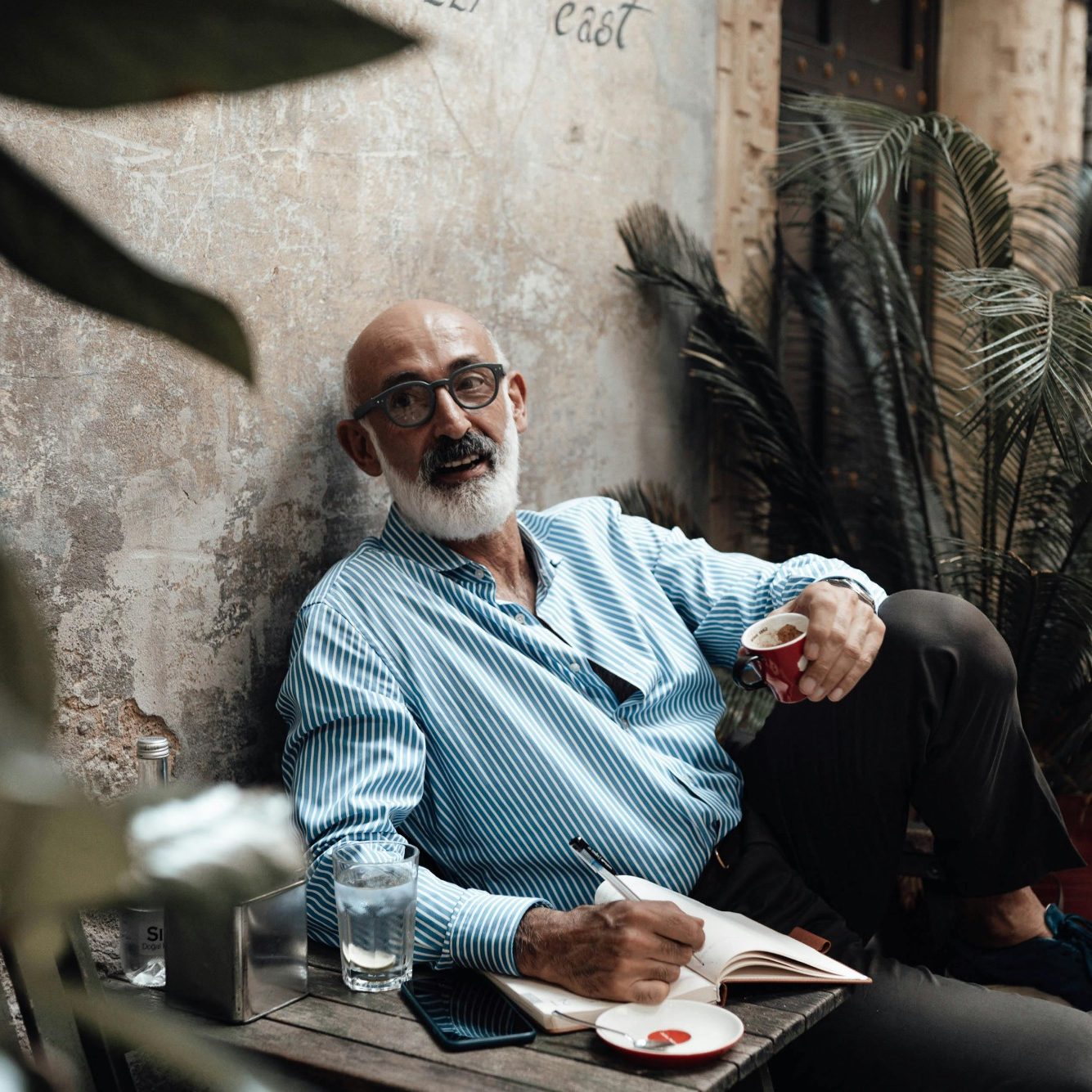 Elderly man in glasses, sitting with a notebook and drink in a rustic setting.