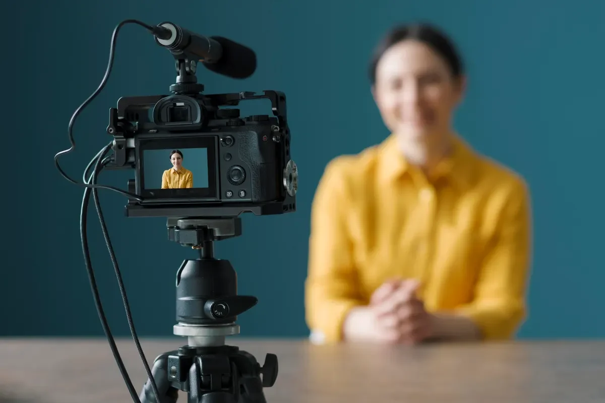 A person in a yellow shirt sits at a table, facing a camera on a tripod.