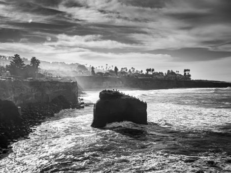 Black and white seascape featuring rocky cliffs and turbulent waves under a cloudy sky.