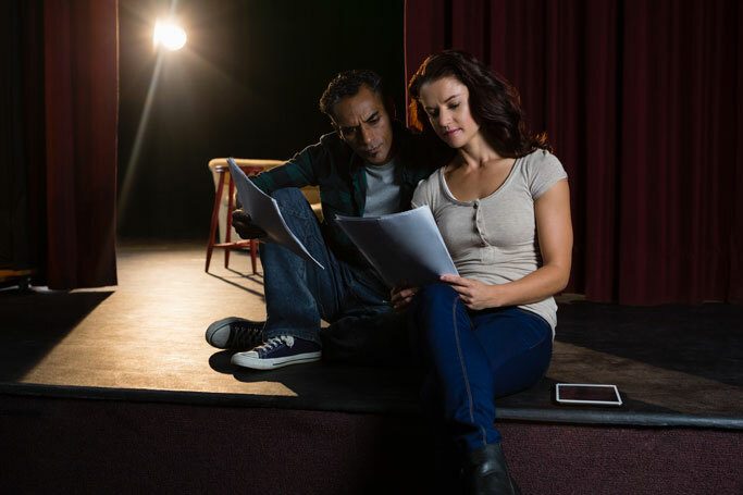 Two actors rehearsing lines together on stage with a spotlight in the background.