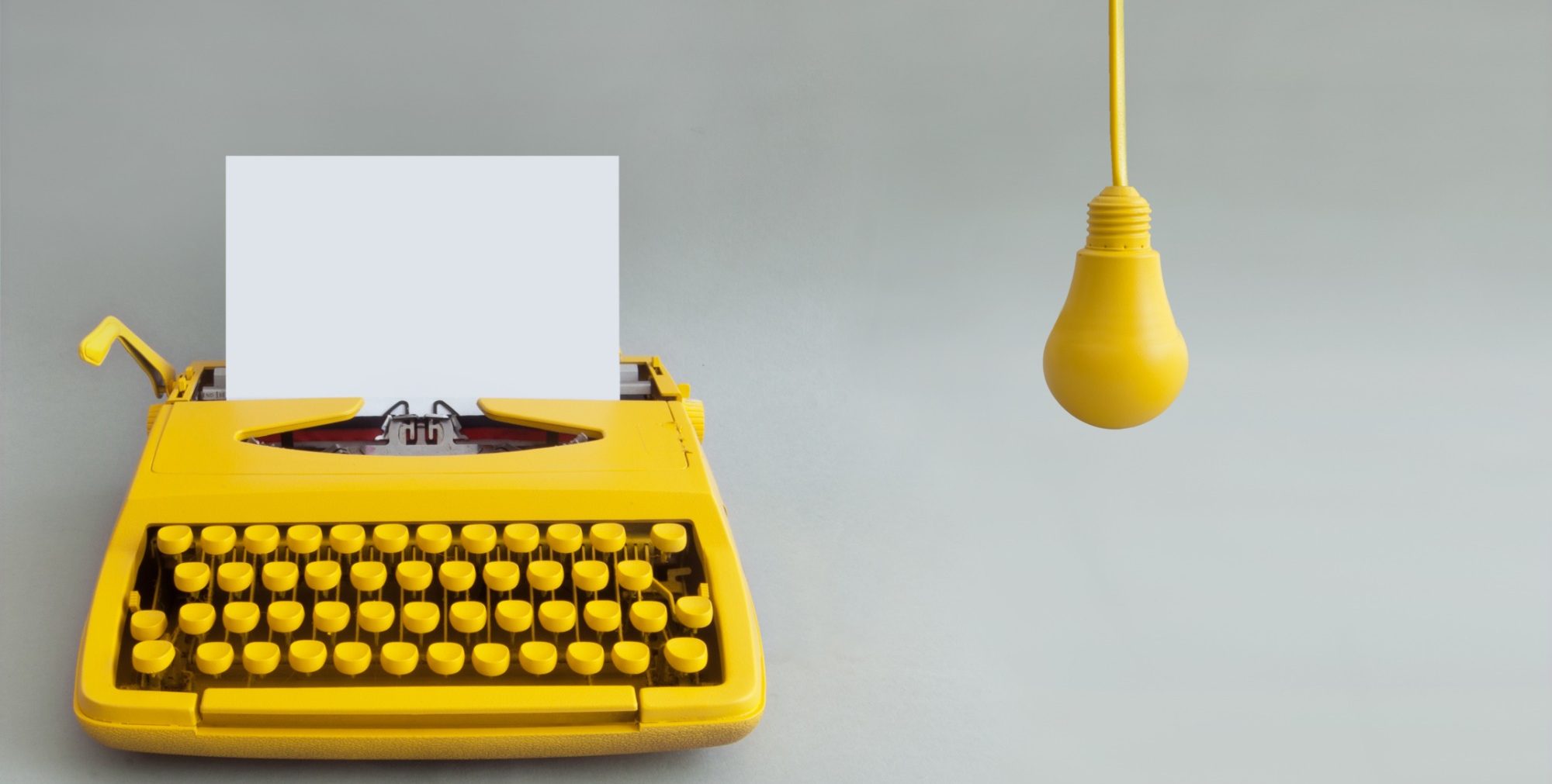 Yellow typewriter with a blank sheet of paper next to a hanging yellow light bulb.