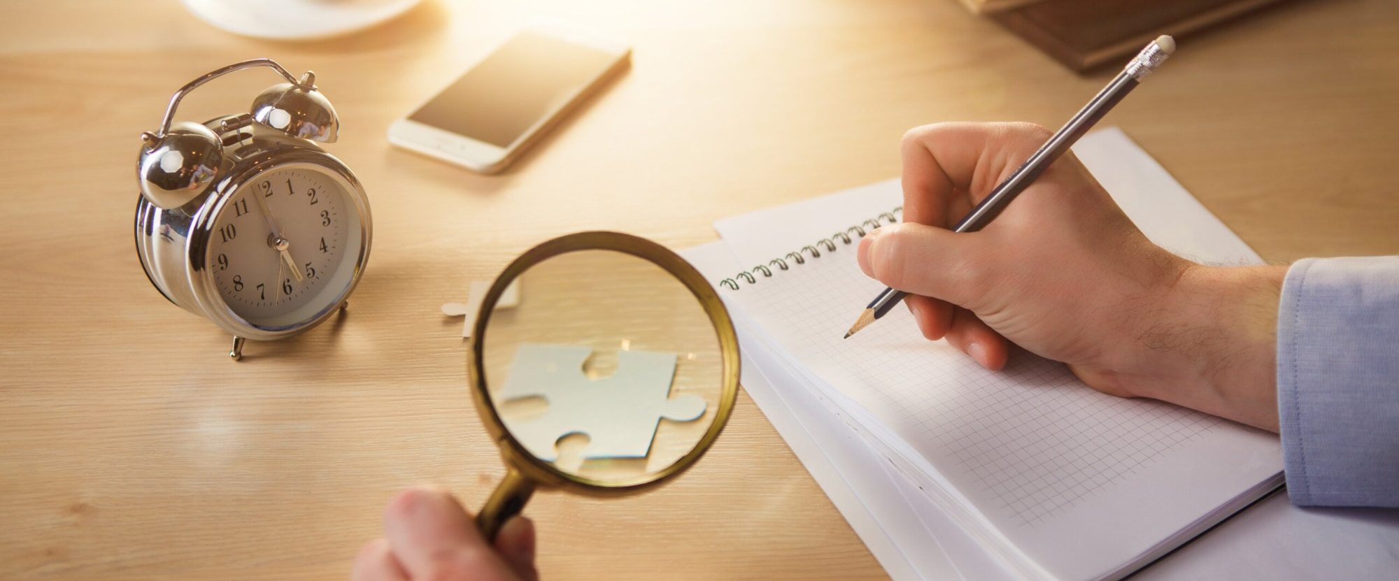 A hand holds a pen over a notebook, with a magnifying glass and alarm clock nearby.