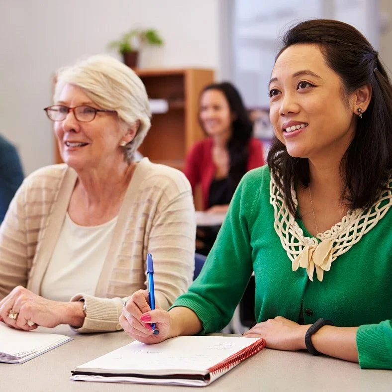Two women sit in a classroom, engaged in a discussion, with notebooks and pens in hand.