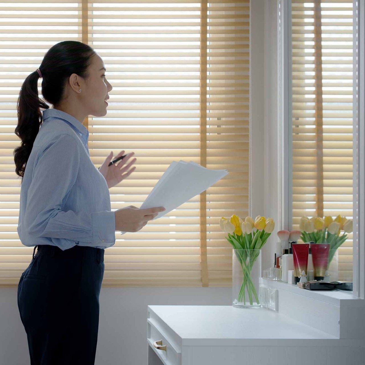 A woman stands by a vanity, holding papers and speaking, with flowers in a vase nearby.