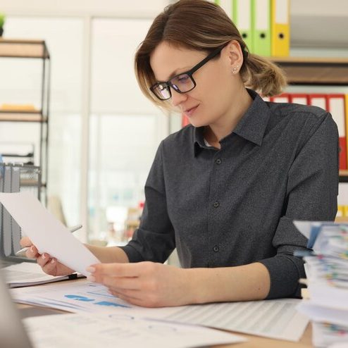 Woman in glasses reviewing documents at a desk with paperwork and office supplies.