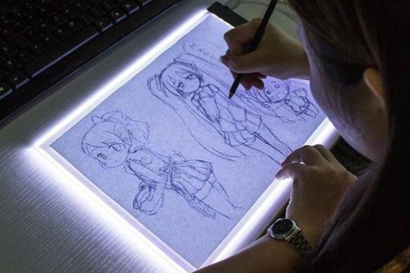 Person sketching anime characters on lightbox with pencil.