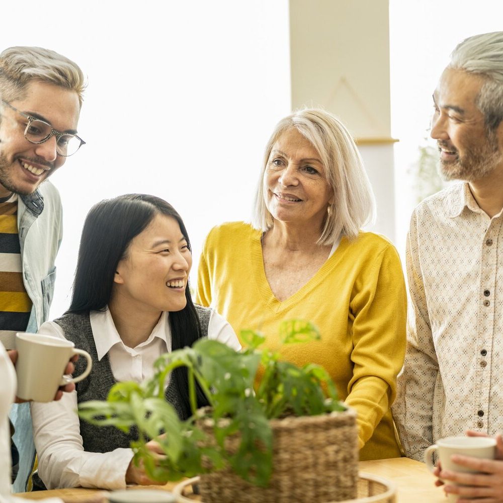 Five people smiling and engaging around a table with plants and coffee cups.
