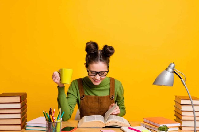 Person with glasses holding a yellow mug, reading a book, in front of a bright yellow wall.