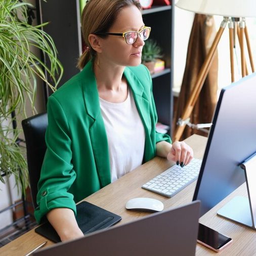 Woman in a green blazer working at a desk with multiple computer monitors.