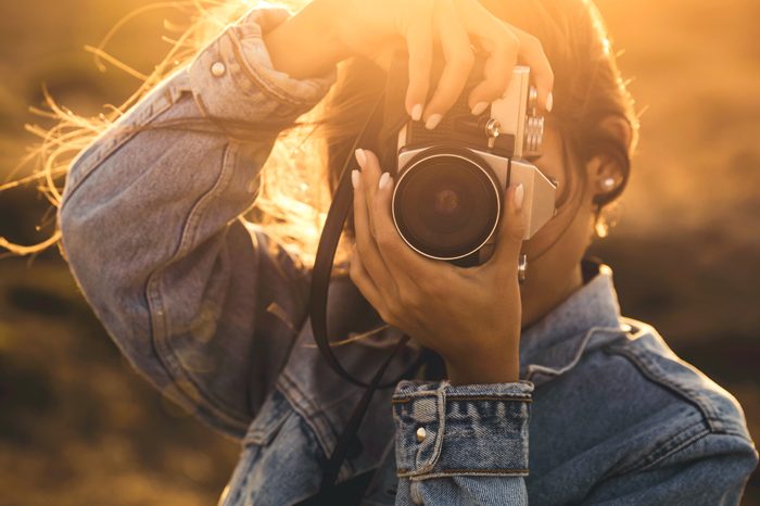 A person in a denim jacket holds a camera, capturing a sunset scene.