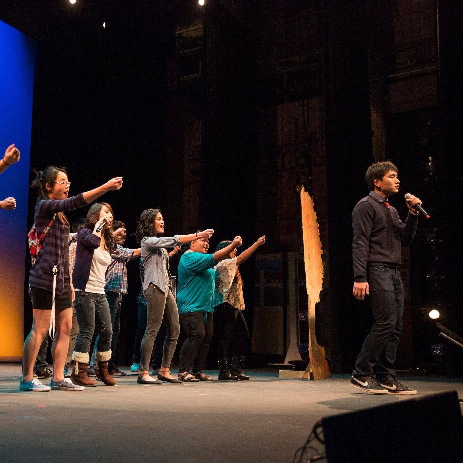 A group of people on stage performing, with one person speaking into a microphone.