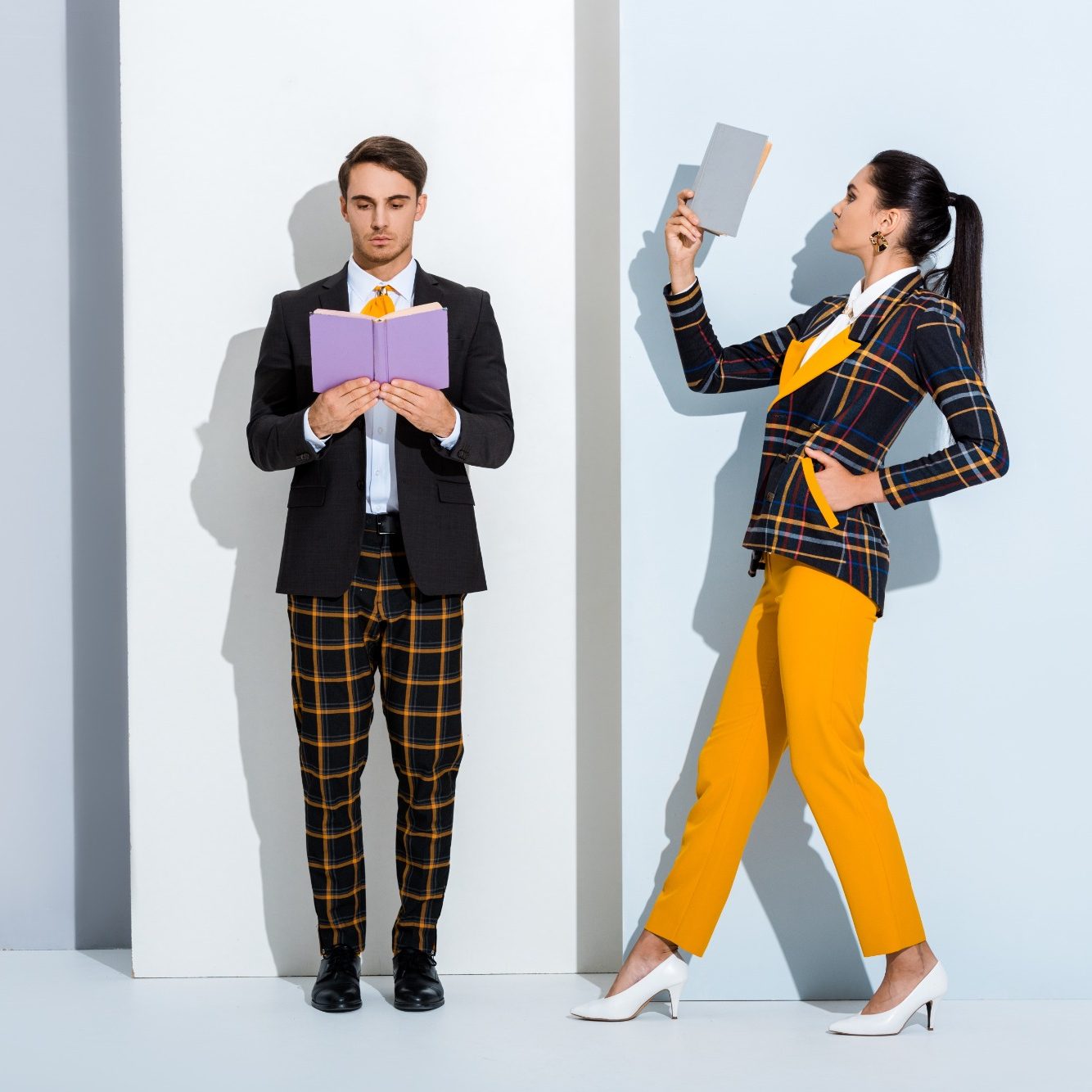 A man in a black suit and a woman in an orange plaid outfit pose against a light background.