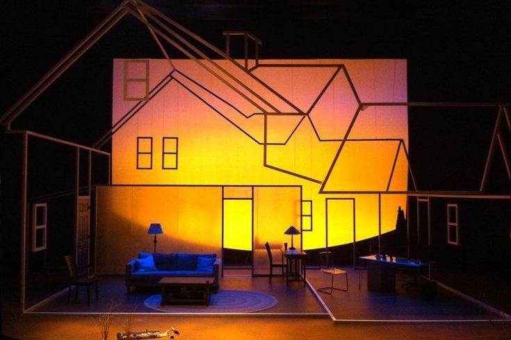 Stage set depicting a simplified house outline with warm lighting and minimal furniture.