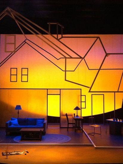 Stage set featuring a minimalist house design with warm lighting and simple furniture.