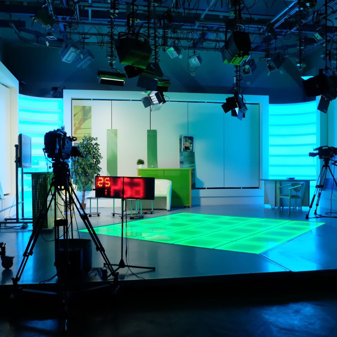 Modern TV studio with green accent lighting, cameras, and a digital clock display.
