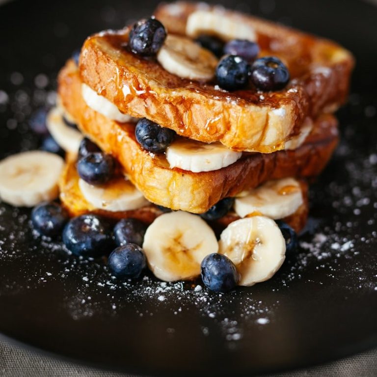 Stack of French toast topped with banana slices, blueberries, and syrup on a black plate.