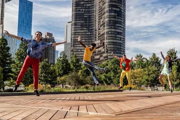 Four people dancing joyfully outdoors with urban buildings in the background.