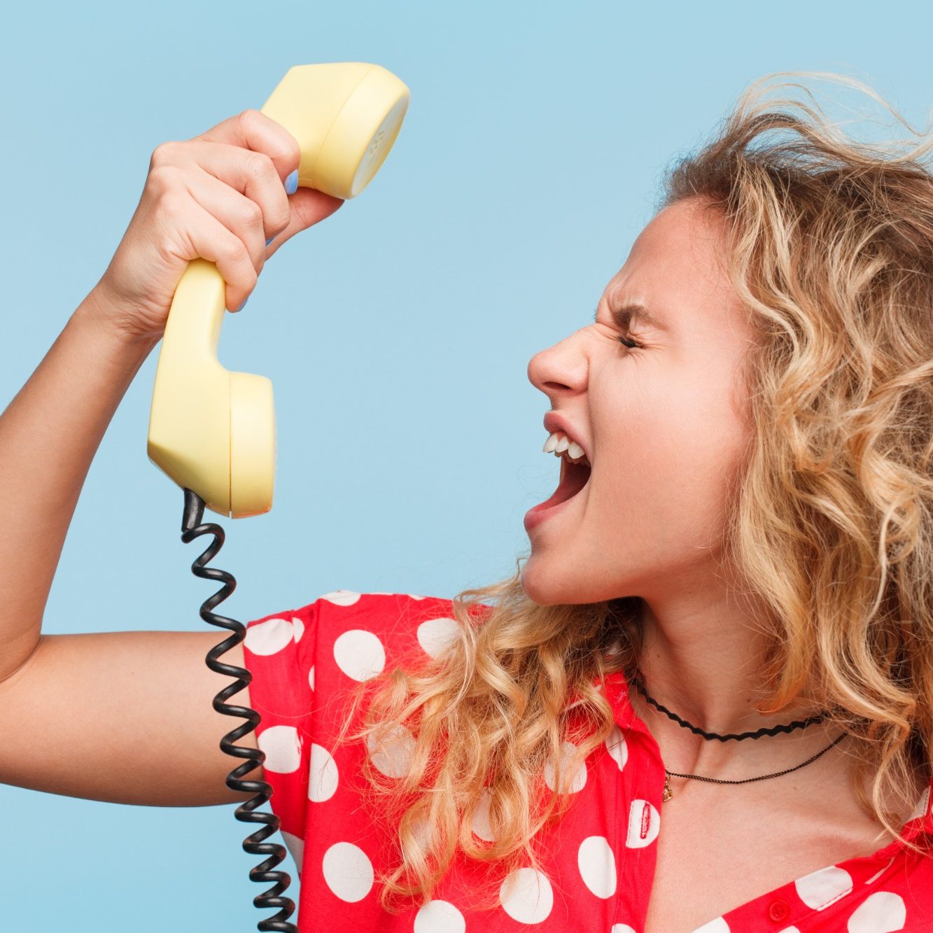 Woman with curly hair in a polka dot shirt shouting into a yellow telephone handset.