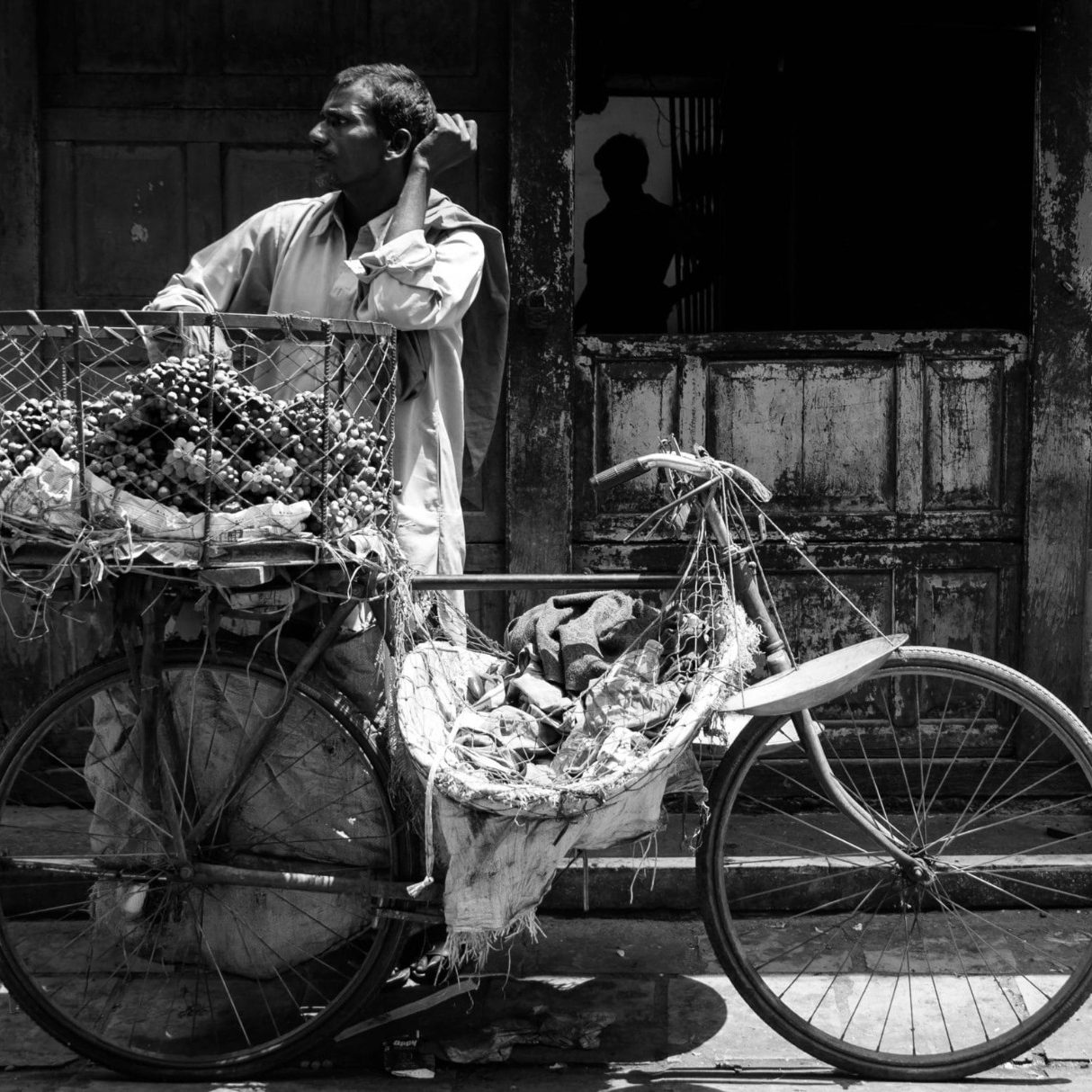 A man standing by a bicycle loaded with goods in a narrow street.