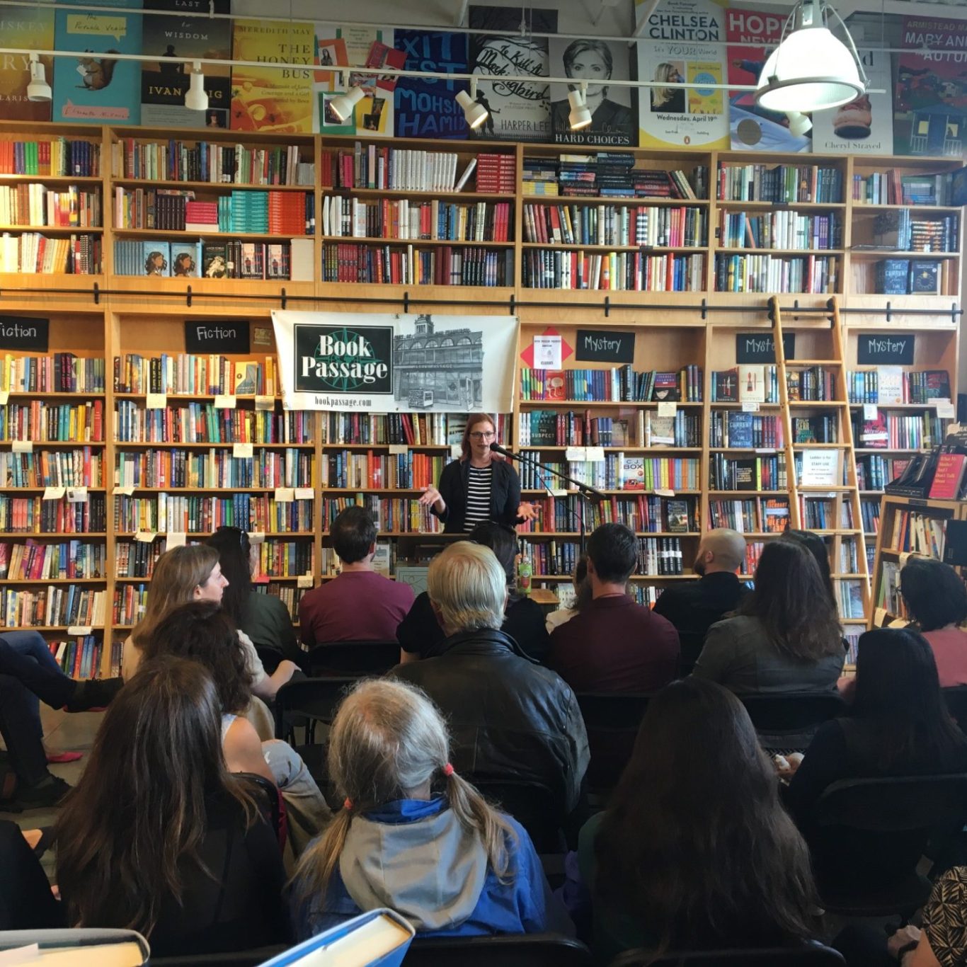 Author speaking to an audience in a bookstore surrounded by shelves of books.