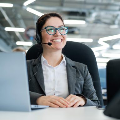 Smiling woman in a headset at a desk with a laptop, in a modern office setting.